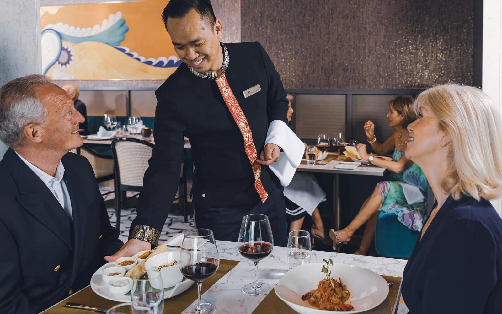 Saga cruise fine-dining photography — waiter serving a pasta course to a smiling couple at their table