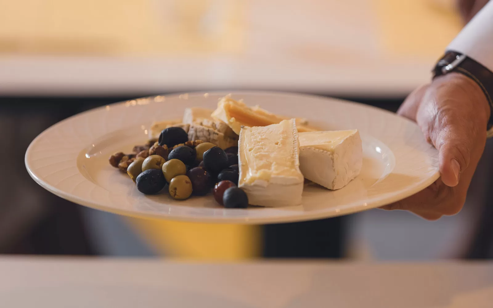 Saga cruise fine-dining photography — cheese-board plate with brie, olives and walnuts held by a waiter